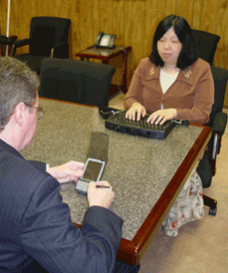 A deaf-blind woman using a device with a keyboard and Braille output sitting in front of a man using a device to communicate. 