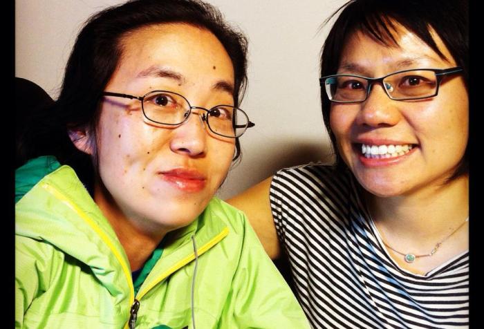 Two Asian American sitting side-by-side. The woman on the left is in a wheelchair; she is wearing a green rain jacket and glasses. The woman on the right has short black hair, glasses and is wearing a striped-t-shirt. They are both smiling at the camera.