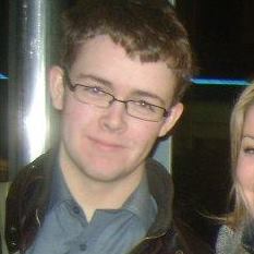 Young white man with short sandy brown hair. He is wearing glasses and a blue button-down shirt. Over his shirt, he has a brown leather jacket. To the right of the image, a face of a white woman with blonde hair is partially cut off from the photo.
