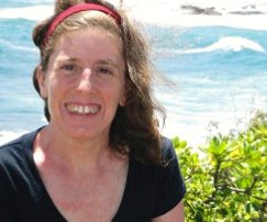 Photo of a white woman smiling at the camera wearing a short-sleeved black t-shirt. The background is the ocean and some green bushes.