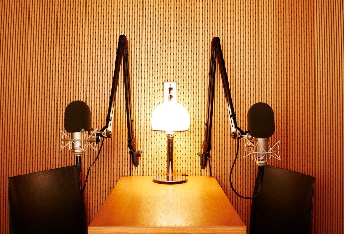 Picture of a small wooden table with two black chairs, one on the left and one on the right. At the center of the table is a small lamp and two swinging arms holding microphones for recording.
