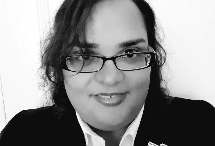 Black and white frontal headshot picture of a Latina woman in her mid to late 20s. She is smiling, has eyeglasses and is wearing a white shirt with a black blazer. In her lapel she is wearing a pin with the logo of the National LGBTQ Task Force, and a brooch in the shape of an owl.