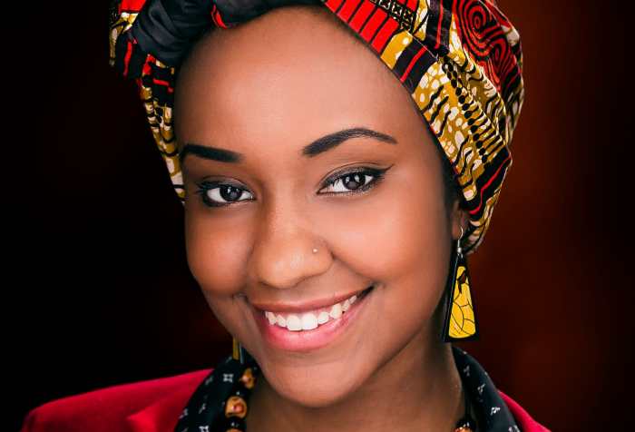 Image description by Azza: Azza, a Black woman, wears a bright red and gold hijab with an East African print, red blazer, and black button down blouse. She is smiling at the camera.