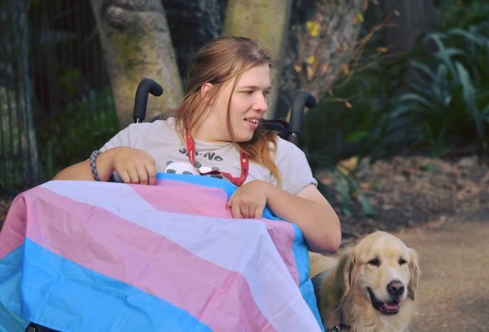 Photo of Annabelle Oxley, a young white woman draped in a trans flag and her assistance dog Jasper, a golden retriever. 