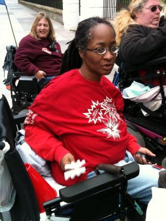 An African-American woman wearing eyeglasses and dressed in a long-sleeved red shirt. She is sitting in a wheelchair.