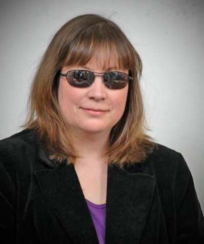 Middle aged white woman with shoulder-length brown hair. She has sunglasses on with reflective lenses and a black blazer and purple t-shirt