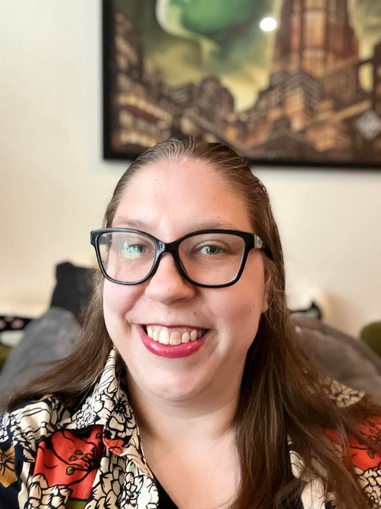 Photo of Anna, a white nonbinary person with dark blond hair and glasses, smiling while sitting on a couch. They wear a black v-neck t-shirt and a floral print button down shirt.