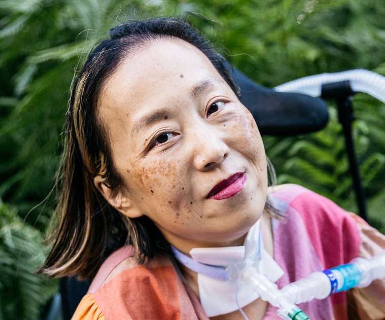 Alice Wong, Asian American woman in a wheelchair with a tracheostomy at her neck connected to a ventilator. She’s wearing a pink plaid shirt and a magenta lip color. She is smiling and behind her are a bunch of trees. Photo credit: Allison Busch Photography.