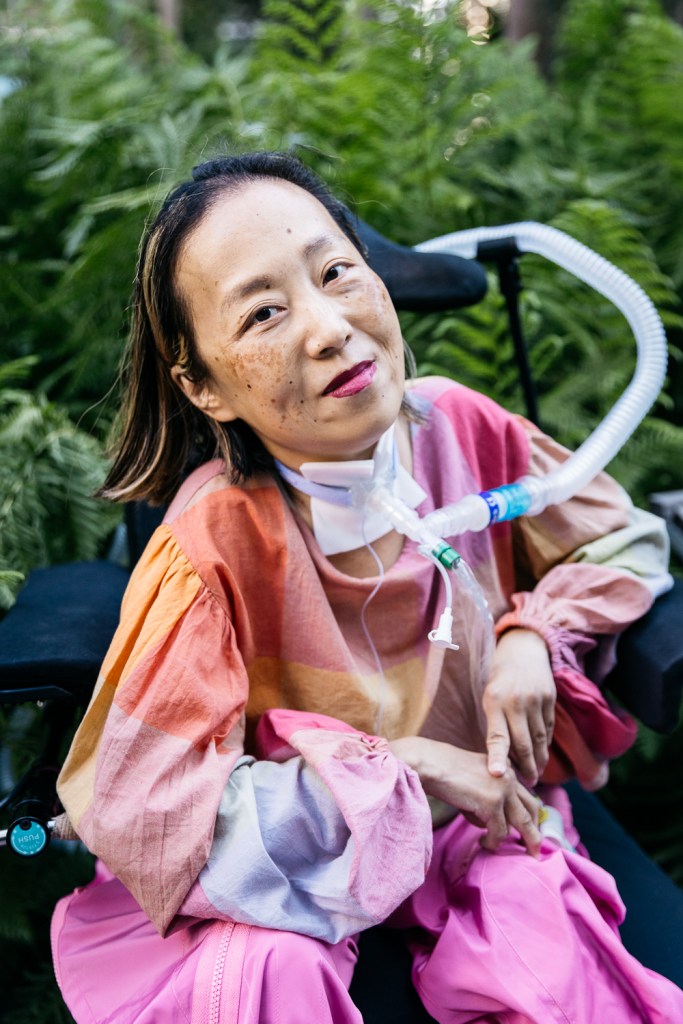 Asian American woman in a wheelchair with a tracheostomy at her neck that has a tube connected to her ventilator. Her head is tilted to the left side, she is smiling, and she is wearing a magenta lip color. Her shirt is plaid with pale pink and other pastel colors and she is wearing pink pants. Behind her is a lot of greenery from bushes and trees at a park. Photo by @allisonbusch_photography