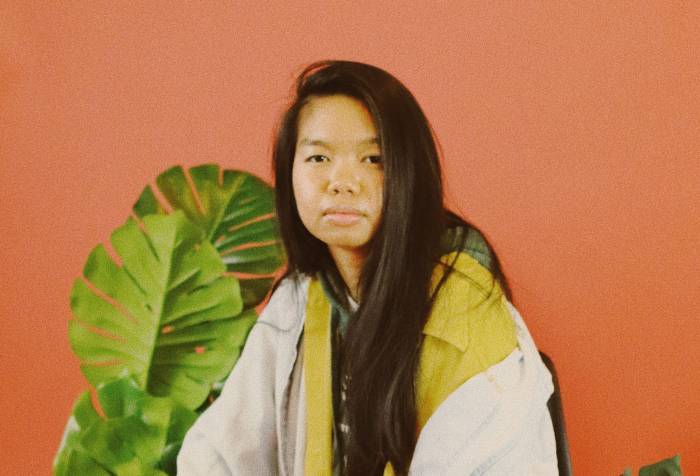 Allison Masangkay (aka DJ Phenohype), a Filipinx femme, sits in a chair wearing an outfit of yellows, greens, lavender, and light wash denim blue with a warm orange backdrop, surrounded by several green plants. They look directly at the camera with a serious expression and their hands resting on their knees. Photo by Bianca Recuenco.