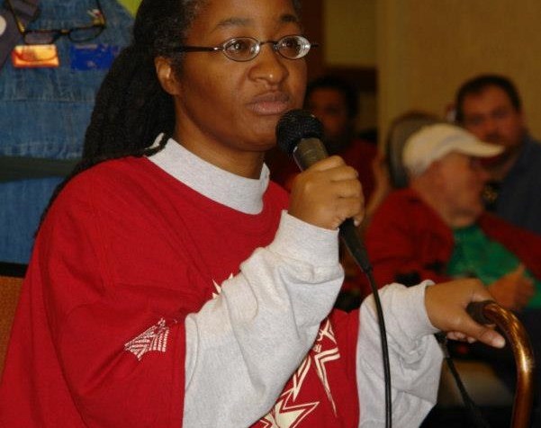 An African-American woman in a long-sleeved red and white shirt. She is wearing eyeglasses and holding a microphone at a meeting.