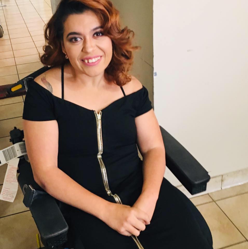 A photo of Ashley, olive-skinned Filipina/White woman with curled reddish brown hair down to her shoulders, smiling with her head slightly to the side. She is sitting in a power wheelchair wearing a black cocktail dress with a gold zipper going down the middle with her hands crossed in her lap