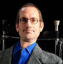 A middle-aged white man with brown hair and glasses. He is sitting in a wheelchair and wearing a dark gray jacket and a blue shirt.