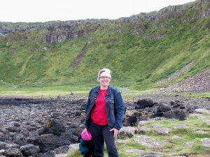 A white woman dressed casually standing at a valley with green hills and black rocks in the background.