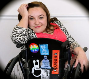 Image of a woman in a wheelchair with her hand cradling her head. She has long brown hair and the back of her wheelchair has various stickers denoting disability and queer culture