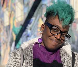 A prince wearing a purple sweater with green hair smirks at the camera.