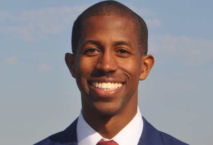 Dr. Justin Bullock, a Black man with short hair wearing a navy suit with a white shirt and dark red tie. He is smiling at the camera.