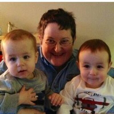 A woman with short hair and glasses holding twin boys who are all smiling at the camera.