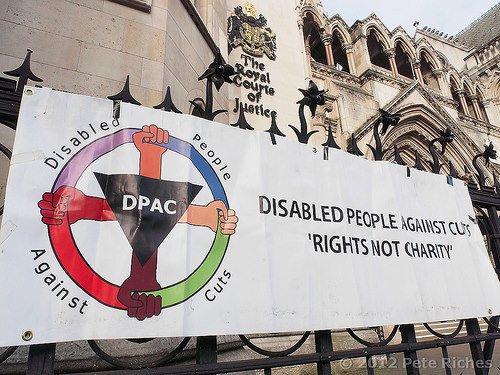 Banner outside of the Royal Court of Justice in the United Kingdom. The banner has a circular logo with 4 fists positioned in the north, west, east, south position. The text reads: DPAC Disabled People Against Cuts. "Rights, Not Charity."