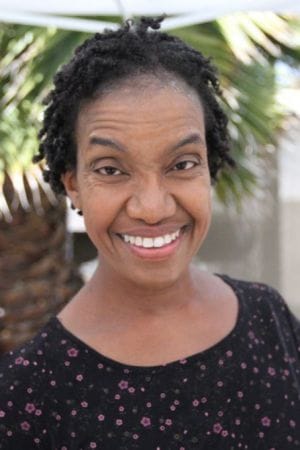 Diana Elizabeth Jordan, a Black woman with short hair in twists smiling at the camera. She is wearing a black scoop neck shirt with a floral print.