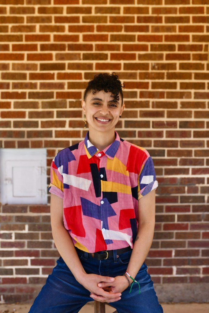 dena, with short black hair and shaved sides, sitting on a stool and smiling against a brick wall wearing a pink shirt with blue, red, white, and black rectangular shapes. They have their hands folded in front of them and are wearing blue jeans and a red, black, green and white bracelet.