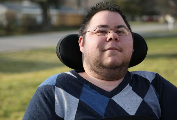Dominick Evans, a nonbinary trans masculine person looks at the camera, with a slight smile on his face. His eyes are also smiling. He has short, dark spiky hair, and wears glasses. He has stubble along his jaw, cheeks, and chin. He is wearing a argyle sweater with various shades of blue, gray, and black. His wheelchair headrest is visible behind his head, and there is green grass and the neighborhood behind him, faded in the background.
