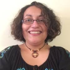 Light brown Puerto Rican woman in her 30s wearing a black top. She is wearing glasses and a brown circular necklace. She is smiling and has curly shoulder-length hair. She is wearing glasses and a brown circular necklace.