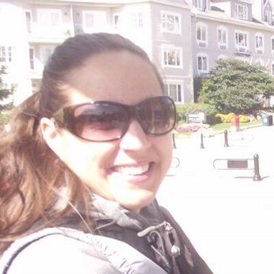 A white woman with long brown hair in a pony tail. She is wearing sunglasses and smiling at the camera.