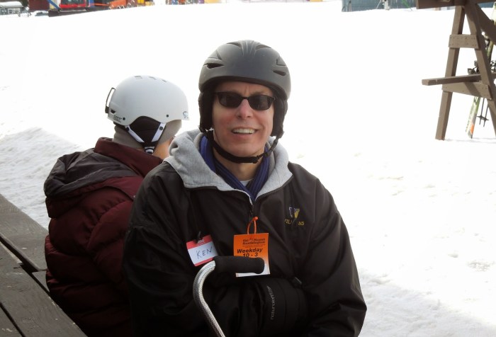 White man in a black ski jacket. He is wearing a helmet against a snowy background. Another person in a helmet is sitting behind him.