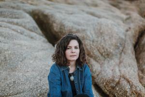 A young white woman with curly long brown hair sitting in front of large rocks wearing a blue denim shirt with a large round pendant around her neck.