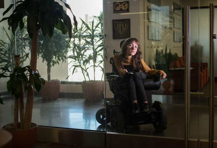 Emily sits among plants in a power wheelchair behind a glass door. She is dimly lit, with long brown hair, a tan turtleneck, and black pants and boots.