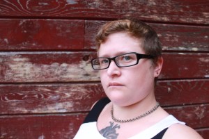 Image description: A head and shoulders shot of s.e. smith, a white person with short brown hair and glasses, posing against the wall of a red building. 