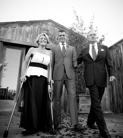 Black and white outdoor image with a barn in the background. A white woman in her 60s is wearing a white and black sleeveless dress. She is using crutches. She is standing left. In the center is a young man, her son dressed in a gray suit. On the right is an older white man in a dark business suit.