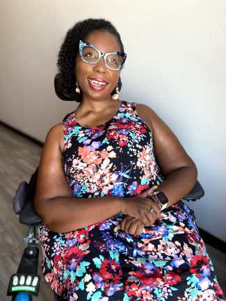 Black woman in a power chair wearing a sleeveless dress with a floral print in bright colors. Her eyeglass frame is cat-eye shaped black and teal. She is smiling and looking slightly away from the camera.