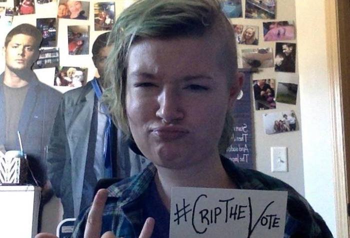 Image of a young white woman with blonde hair combed to the right side of her face, the left side of her hair is shaved off. Behind her are photos and poster typical of any college dorm room or bedroom. Her eyes are squinting and her lips are pursed as her right hand is making a "rock on" pose with her middle and ring finger downward while the others are standing up. On the left hand she is holding a white index card that reads, #CripTheVote.