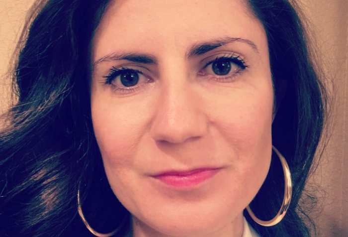 Conchita Hernandez Legoretta, a Latina woman smiling at the camera. She has long wavy brown hair and is wearing gold hoop earrings.