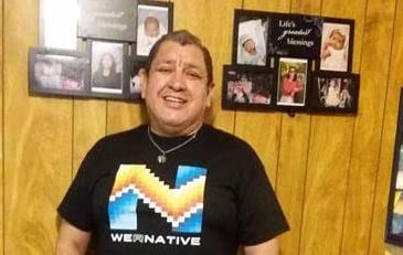 Picture of Hector standing in the hallway of his house. The walls and floors are wooden and there are some pictures of his family on the wall. He is standing and is wearing a T-shirt that reads WeRNative that has an native patterns. He is wearing dark blue jeans and great Crocks. His hands are in his pant pockets and he is wearing a lime green bracelet that reads “Each Mind Matters”. He is smiling at the camera and is wearing a turquoise and silver pendant on a small chain around his neck.