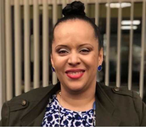 Light-complected Black woman shown shoulders up wearing hair in bun atop her head, blue button earrings, makeup with red lipstick and smiling. She is wearing olive-colored blazer and blue and white patterned blouse with long necklace of various blue-colored pendants