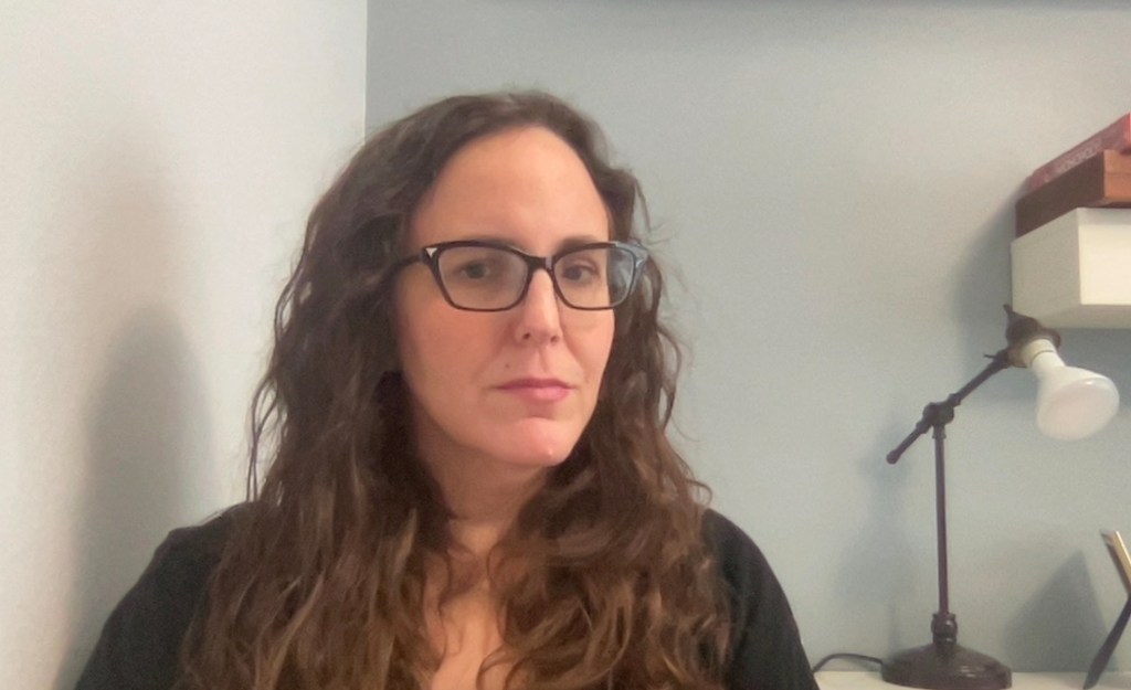 headshot of Justine Barron, a white woman with long wavy brown hair and black glasses, seated in front of a desk lamp.