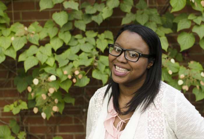 Black woman with black hair and black glasses is wearing a white shirt and smiling in front of a green flowered wall. Photo credit: Reid Beels