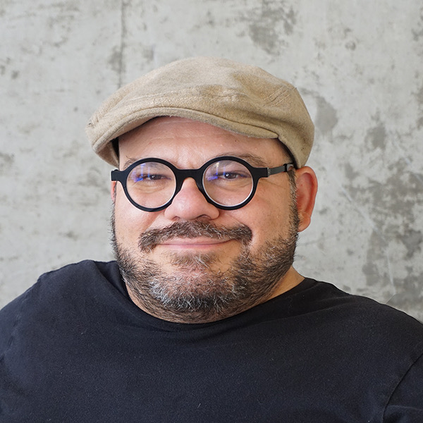 A photo of Joe’s head and shoulders on a gray background. He is a white man with a short cropped, dark beard and is wearing black rimmed round glasses, a black t-shirt, and tan colored flat cap.