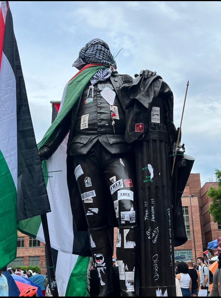 A statue of George Washington in Washington, DC. The statue's face is wrapped in a kuffiyah, it is wearing a Palestinian flag as a cape, and is holding another Palestinian flag. There are various stickers on the chest and body.]