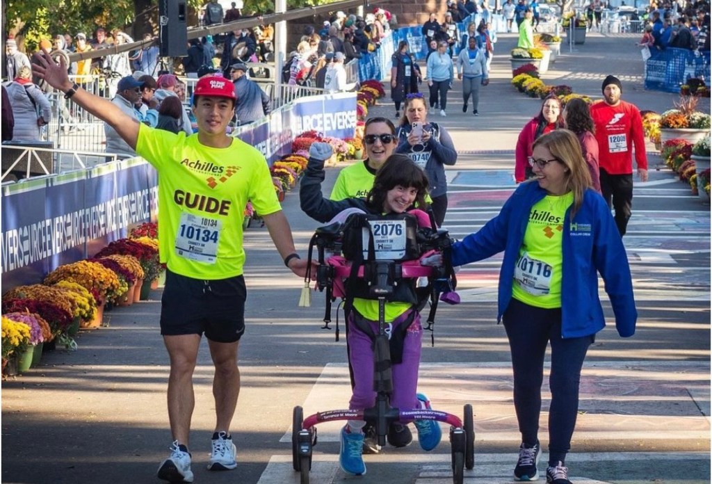 Margaret smiling with her fist held high as she crosses the finish in her pink walker. She has shoulder-length brown hair and is wearing a neon yellow Achilles jersey with purple pants, a black jacket, powder blue gloves, and royal blue sneakers. To the right is her female Achilles guide with brown hair, glasses, a blue jacket open over a yellow Achilles jersey, and black pants. Holding onto the walker with one hand, she is smiling and looking at Margaret. On the other side of Margaret is a male Achilles guide in a yellow jersey, red baseball cap, and black shorts. One hand grips the side of the walker while the other is waving in the air as he looks forward and smiles. Behind Margaret is another female Achilles guide with a black ponytail, yellow jersey, and sunglasses as she smiles.