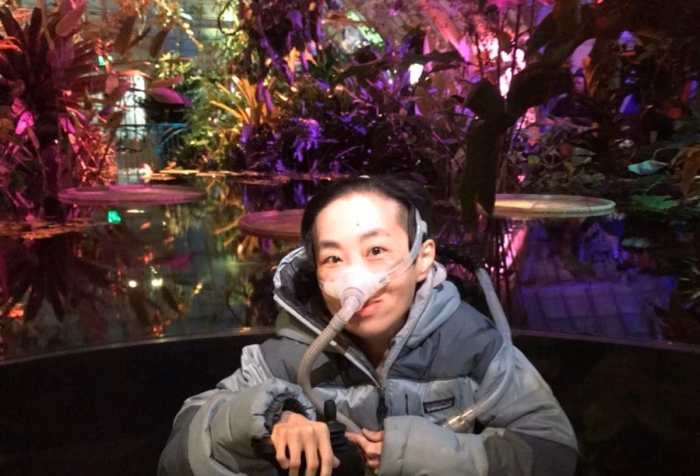 Photo on the of an Asian American disabled woman in a wheelchair in front of a pool of water with tropical water plants in the background lit with purple & magenta lights inside a greenhouse. She is wearing a puffy gray jacket and a mask over her nose attached to a tube.