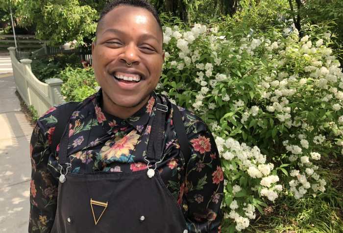 Cyrée Jarelle Johnson, a black transmasculine person with medium brown skin, a septum ring, and a big smile stands in front of a hydrangea bush that is behind a white picket fence on a sunny day. It’s so bright his eyes have squinted closed. He is wearing black shortalls, a gold triangle necklace, and a silky, floral black shirt rolled up to the elbows. Photo by J.D. Stokely