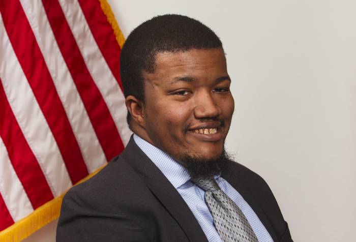 Image of a black man smiling with arms crossed in front of flag