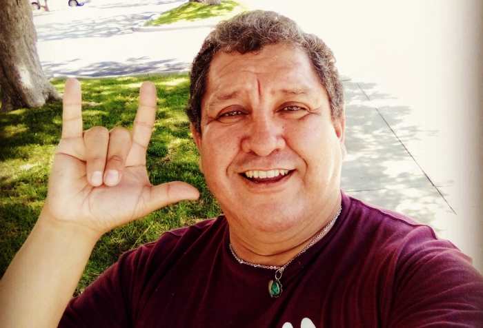 Photo of Héctor M. Ramírez, a Latino Chiricahua Apache man wearing a maroon t-shirt that says ASL with 2 illustrations of hands spelling ‘a’ and ‘f’ as in af meaning “as fuck”. His right hand is showing the sign, "I love you" in American Sign Language.