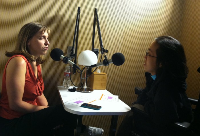 On the left is a white woman with long blonde-brown hair wearing an orange tank top. She is sitting at a table across from an Asian American woman in a wheelchair. She is wearing glasses and wearing a black hoodie. There are microphones on the table and they are having a conversation.