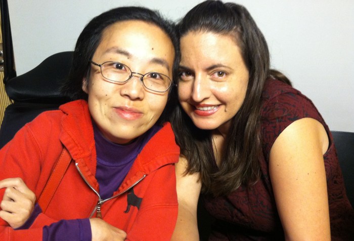 Photo of an Asian woman in a wheelchair w/ glasses wearing a red hoodie. She is on the left-hand side and on the right is a white woman with long brown hair and a magenta short-sleeve top. Both are smiling.
