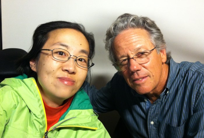 Asian woman sitting on the left-hand side wearing glasses and a green jacket. Next to her is an older white man with curly hair and glasses. He is wearing a blue shirt